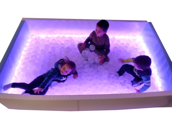 Drei Kinder spielen begeistert in einem LED-beleuchteten Bällebad mit transparenten Bällen und violettem Licht – ein fröhliches sensorisches Erlebnis.