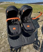 Doppel-Kinderwagen mit schwarzen Sitzen und braunen Griffen am Wasser